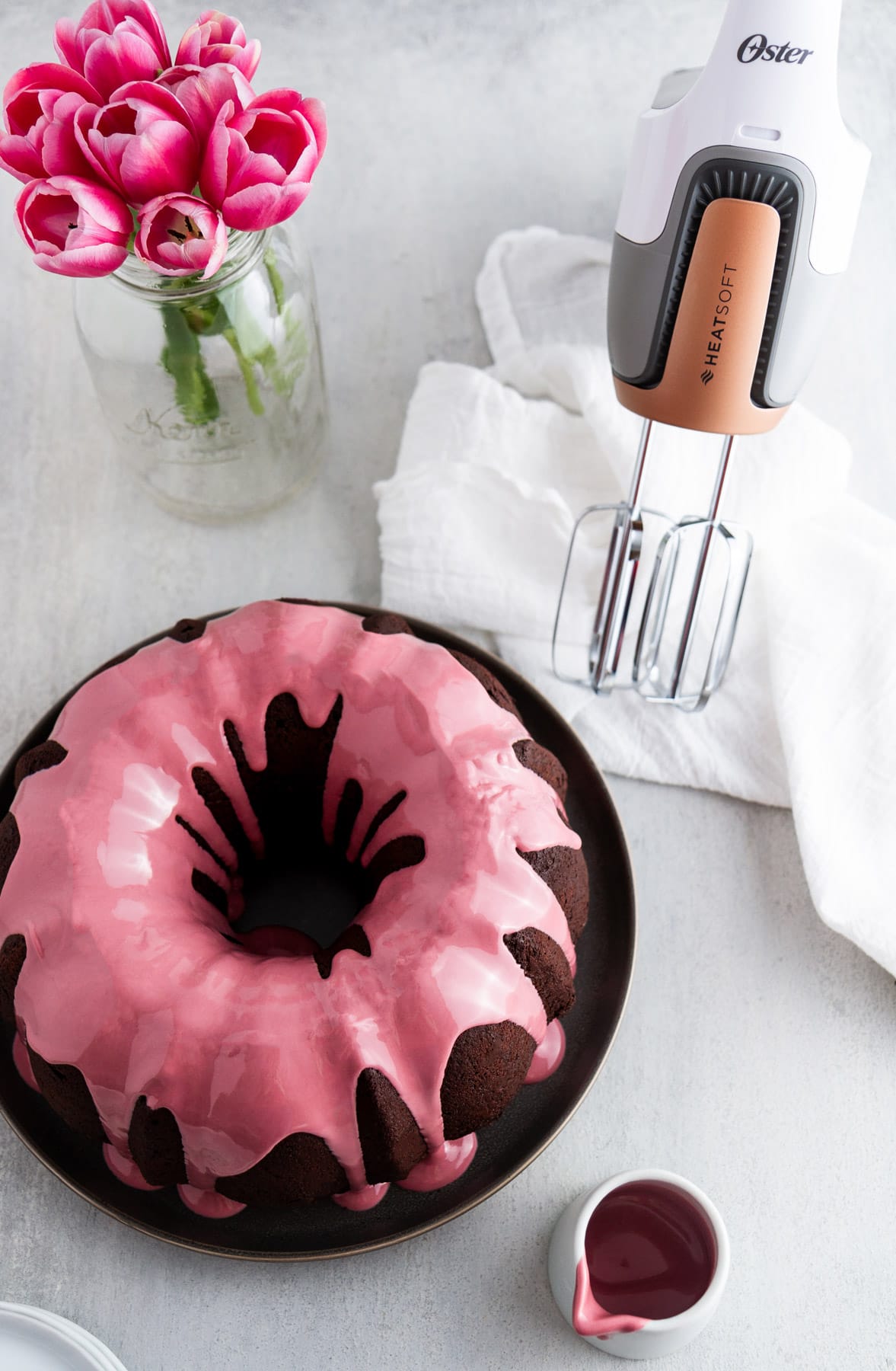 chocolate bundt cake with ruby chocolate glaze on a plate next to a vase of flowers and a hand mixer