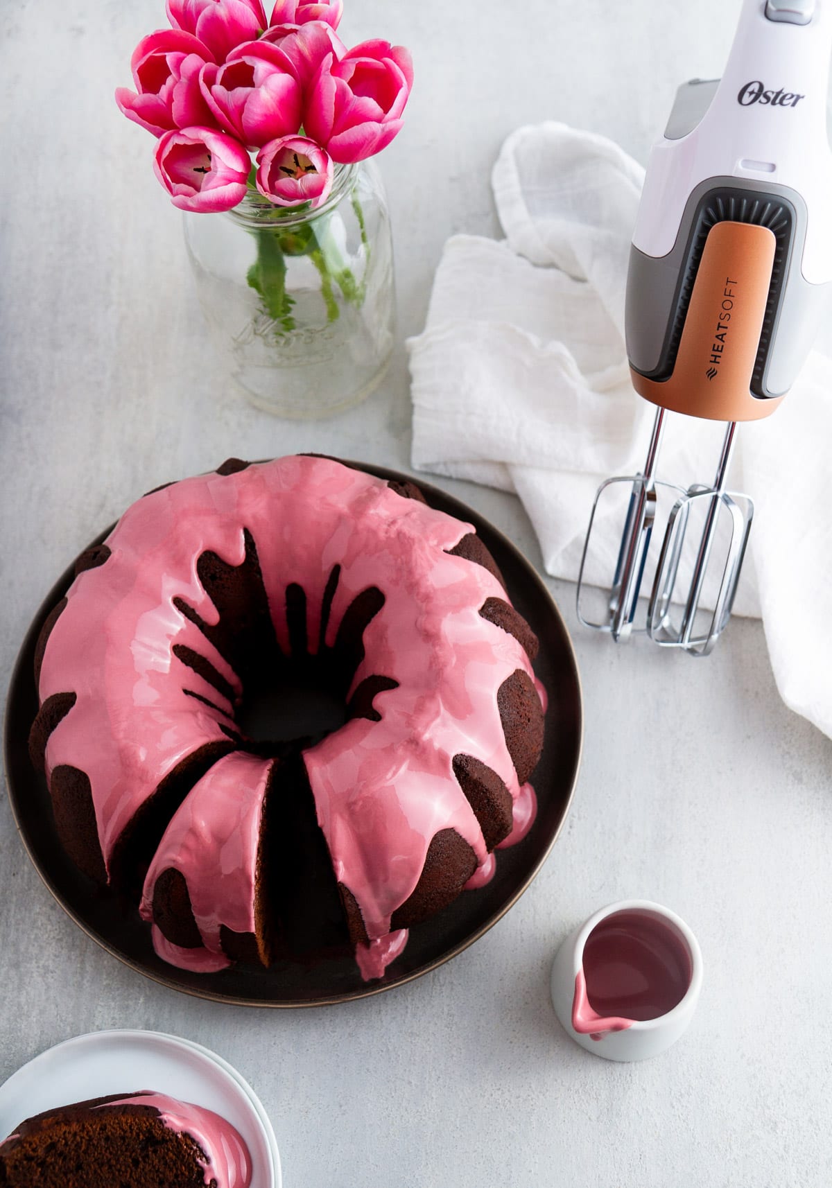 a chocolate bundt cake with ruby chocolate glaze on a plate with a hand mixer and flowers next to it